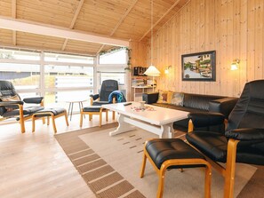 A cozy living room with a wooden ceiling, a large window, a black leather sofa, a white coffee table, and a wooden floor.