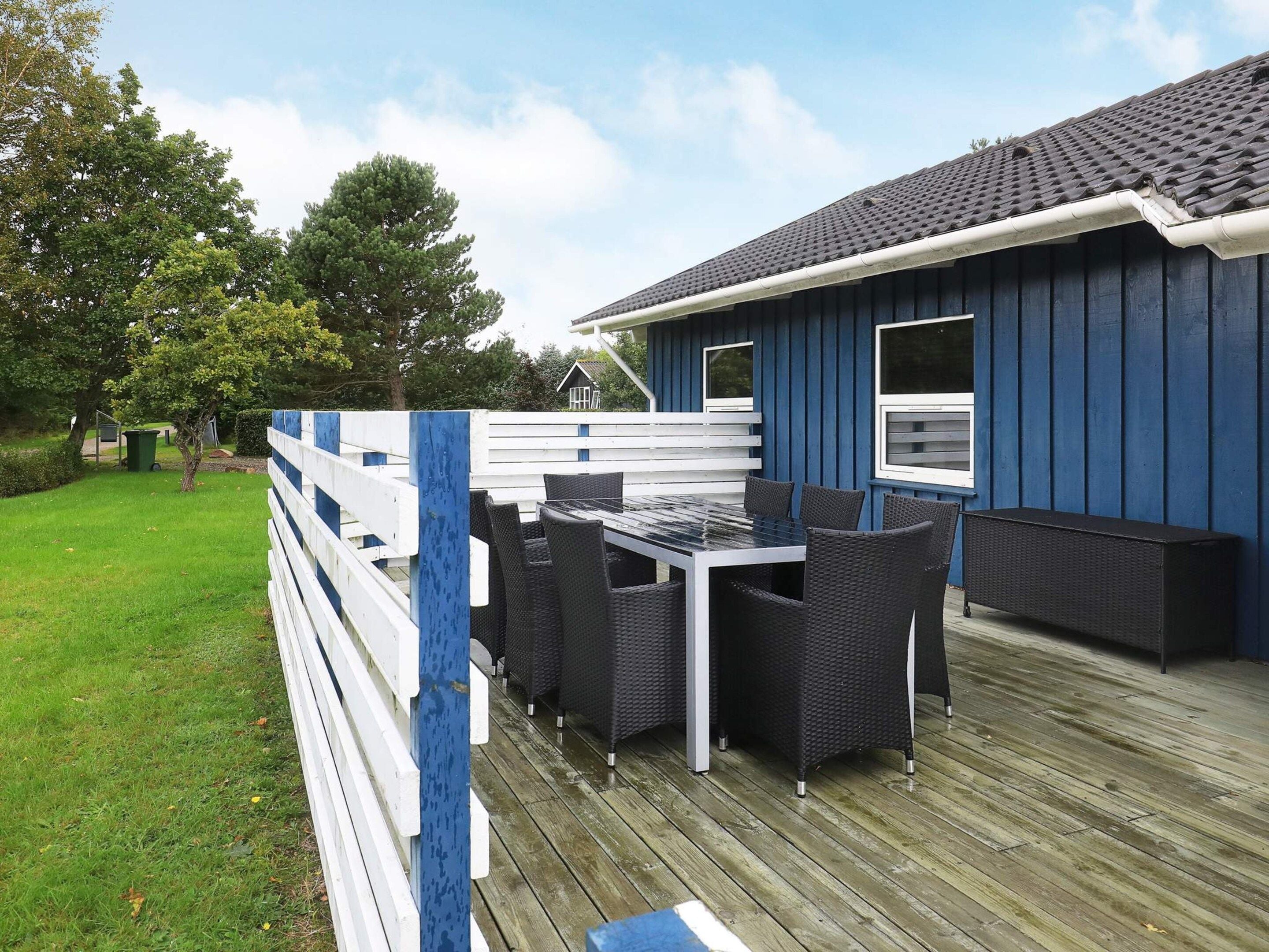 A deck with a table and chairs, a blue building with windows, and a grassy area with trees.