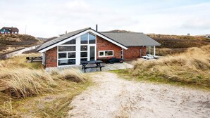A single-story brick house with a large glass door and windows, a covered patio area, and a sandy path leading to it. - 4 Star Holiday Home in Henne (Henne)