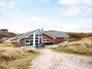A single-story brick house with a large glass door and windows, a covered patio area, and a sandy path leading to it. - 4 Star Holiday Home in Henne (Henne)