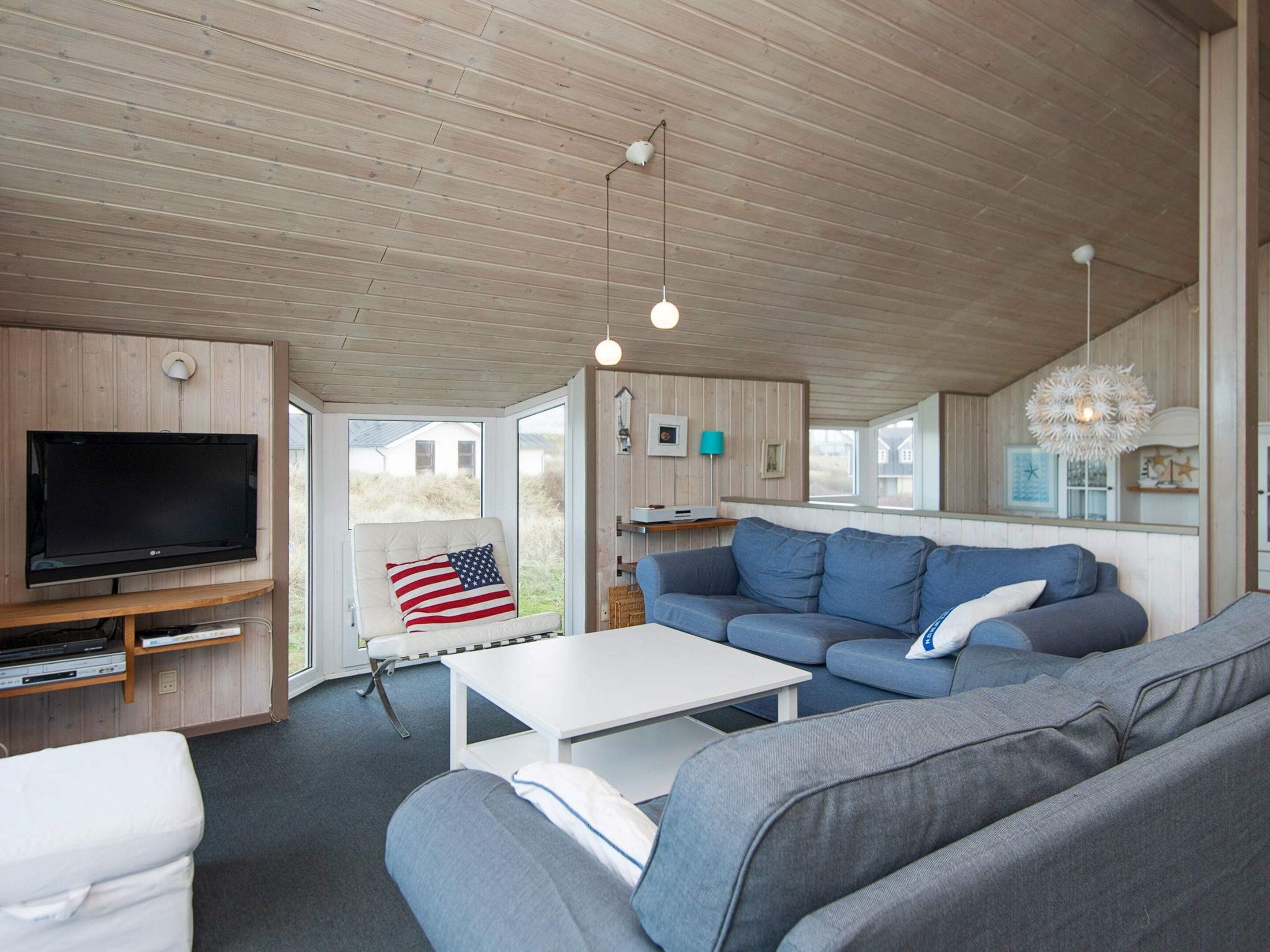 A modern living room with a flat-screen TV, a grey sofa, a white coffee table, and a ceiling with wooden beams.