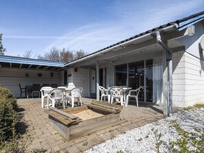 En täckt uteplats med utemöbler, en trälåda för plantering och en byggnad med ett blått tak och stora fönster.