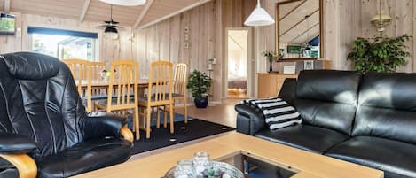 A modern living room with a wooden ceiling, leather sofas, a dining table with chairs, and pendant lighting.