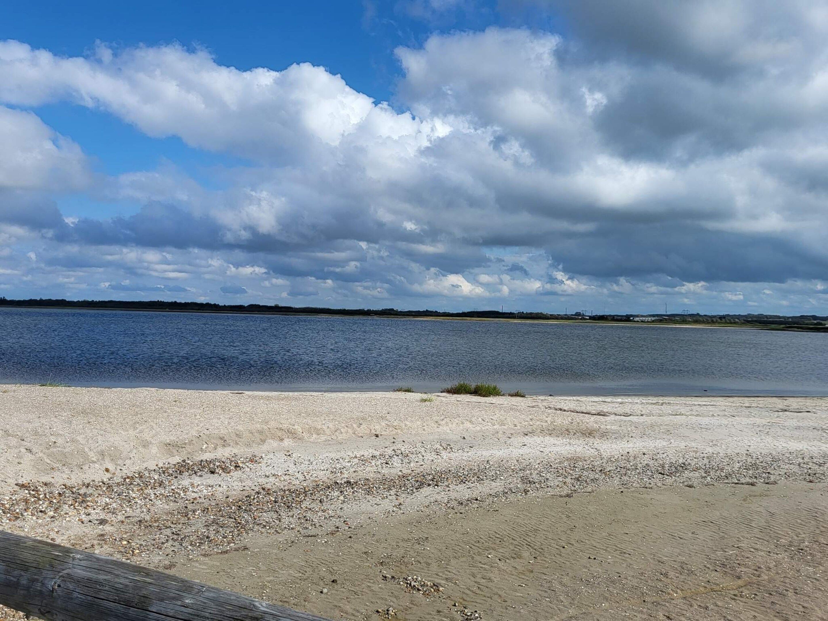 Una playa con un cerco de madera, una orilla arenosa y una masa de agua tranquila bajo un cielo nublado.