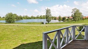 A wooden deck with a railing overlooking a lake, surrounded by greenery and houses in the background. - Holiday Home in Otterndorf (Otterndorf)