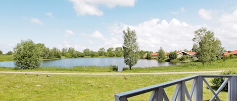 A wooden deck with a railing overlooking a lake, surrounded by greenery and houses in the background.