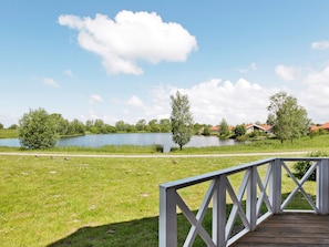 A wooden deck with a railing overlooking a lake, surrounded by greenery and houses in the background. - Holiday Home in Otterndorf (Otterndorf)