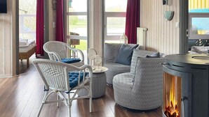 A cozy living room with a sofa, chairs, a patterned ottoman, a fireplace, and a window with curtains. - Holiday Home in Otterndorf (Otterndorf)