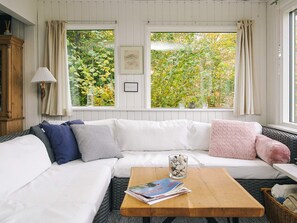 A cozy room with a white sofa, a wooden coffee table, and a view of greenery through large windows. - Lovely Holiday Home in AsnÃ¦s near Sea (Asnaes)
