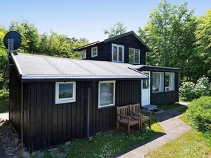 A two-story wooden cabin with a satellite dish, a small porch with a bench, and a garden area with white flowers.