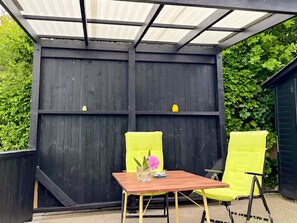 A covered patio with a wooden table and two yellow chairs, a small vase with flowers, and a dark wooden fence. - Lovely Holiday Home in AsnÃ¦s near Sea (Asnaes)