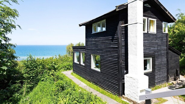 A black wooden house with a white chimney, a clear view of the sea, and lush greenery around. - Lovely Holiday Home in AsnÃ¦s near Sea (Asnaes)