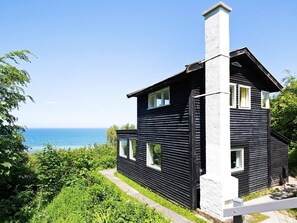 A black wooden house with a white chimney, a clear view of the sea, and lush greenery around.