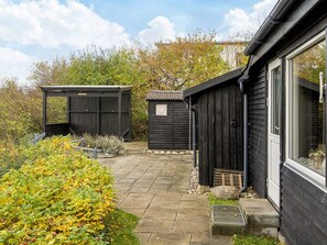 A garden with two wooden sheds, a paved pathway, and a house with a large window. - Lovely Holiday Home in AsnÃ¦s near Sea (Asnaes)