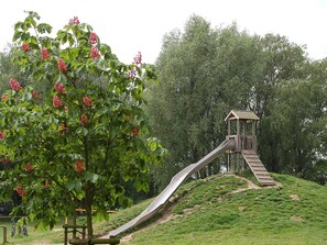 A playground with a slide and a wooden structure, surrounded by green grass and trees. - Holiday Home in Otterndorf (Otterndorf)