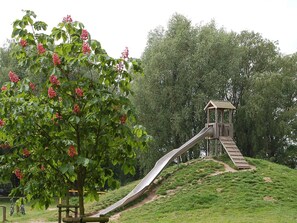 A playground with a slide and a wooden structure, surrounded by green grass and trees. - Holiday Home in Otterndorf (Otterndorf)