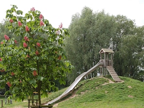 A playground with a slide and a wooden structure, surrounded by green grass and trees. - Holiday Home in Otterndorf (Otterndorf)