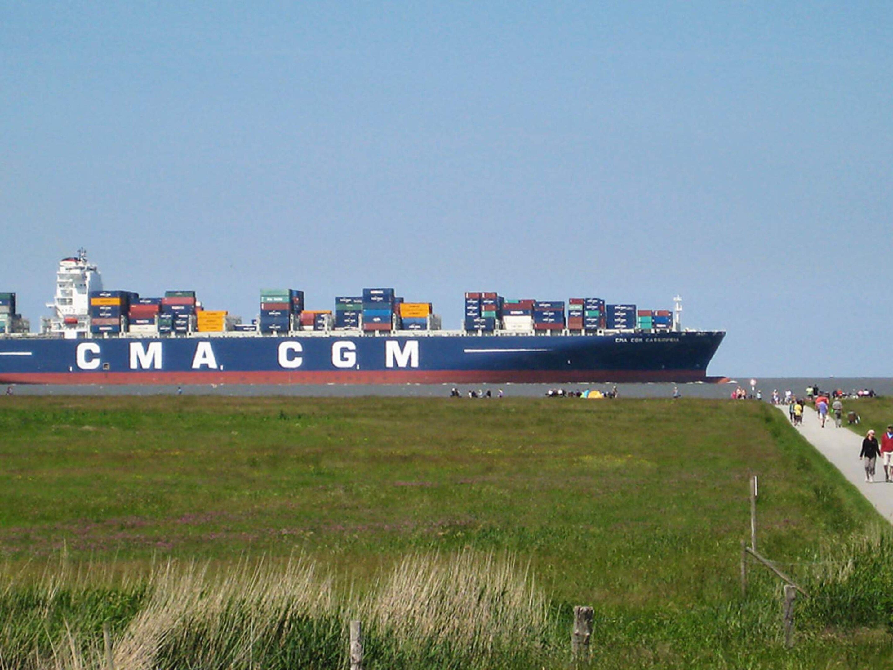 A large cargo ship with the letters 'CMA CGM' on its side, docked at a port with a grassy area and a walking path nearby.