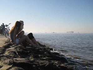 A rocky shoreline with two people sitting on the rocks, a bicycle, and a distant view of the sea with ships. - Beautiful Holiday Home in LÃ¸gstÃ¸r (Otterndorf)