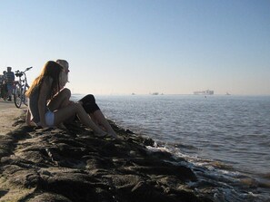 Linha costeira rochosa com duas pessoas sentadas sobre as pedras, uma bicicleta ao fundo, e um mar tranquilo com navios ao longe.