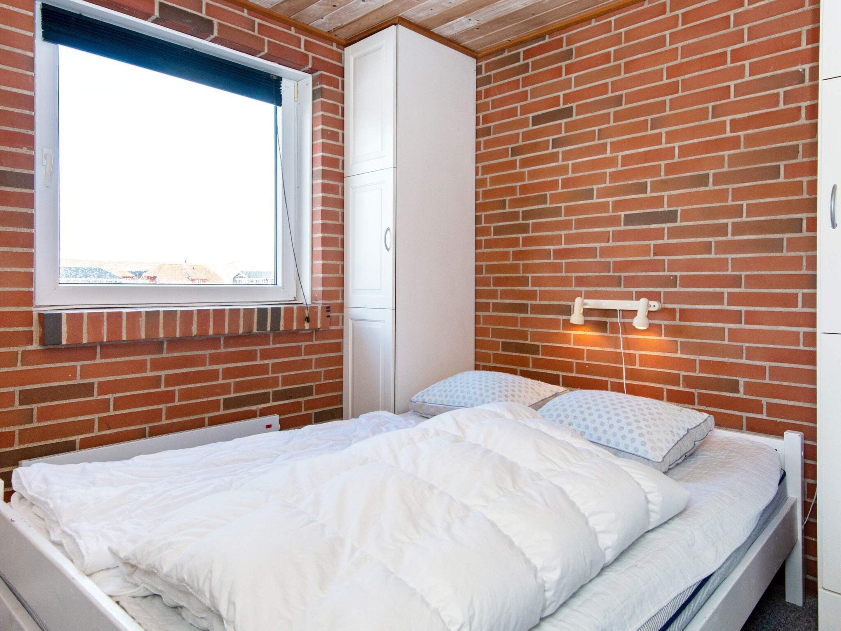 A bedroom with a brick wall, a window, a bed with white bedding, and a wardrobe.