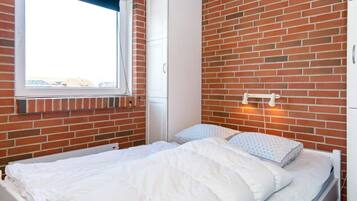 A bedroom with a brick wall, a window, a bed with white bedding, and a wardrobe.