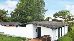 A single-story white building with a dark door, a small porch with a bench, and a paved area in front. - 4 Person Holiday Home in Tranekaer (Tranekaer)