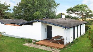 A single-story white building with a dark door, a small porch with a bench, and a paved area in front.