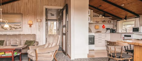 A cozy interior with a wooden ceiling, a sofa, a chair, a dining table, and a kitchen area.