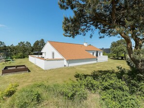 A single-story house with a sloped roof, a fenced yard, and a large tree in the foreground. - 5 Star Holiday Home in Glesborg (Glesborg)