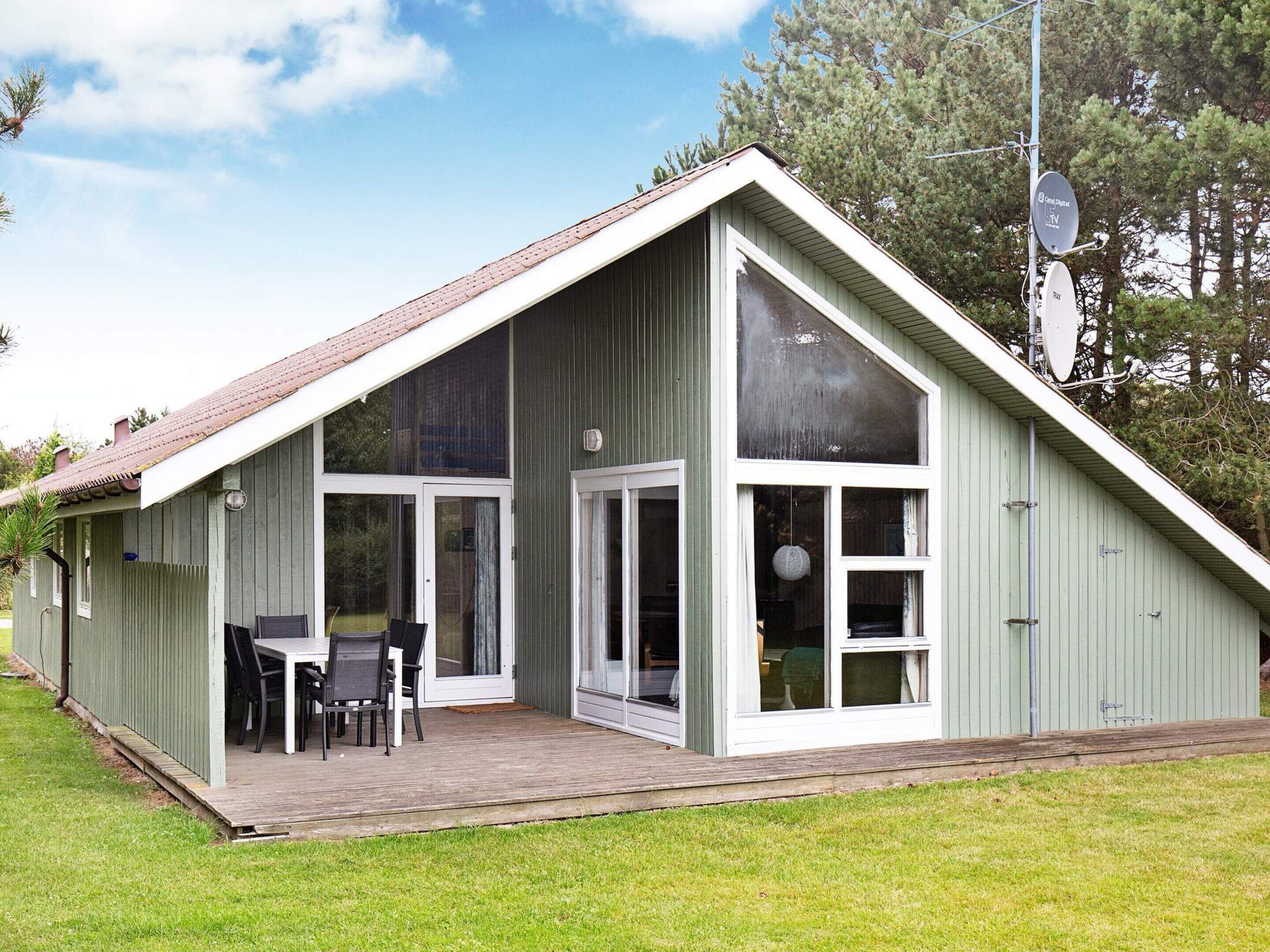 A single-story building with a sloped roof, large glass windows, and a covered patio area with a table and chairs.