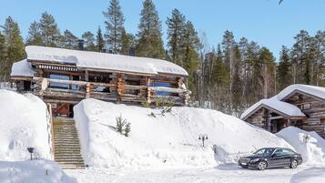 Une cabane en rondins dont le toit est recouvert de neige, avec un escalier menant à l’entrée et une voiture garée devant.