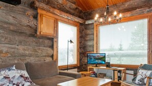 L’intérieur d’un chalet accueillant, avec un plafond en bois, une grande fenêtre munie de stores, un téléviseur, un bureau avec un ordinateur, un canapé et un fauteuil agrémenté d’un coussin à motif de cerf.