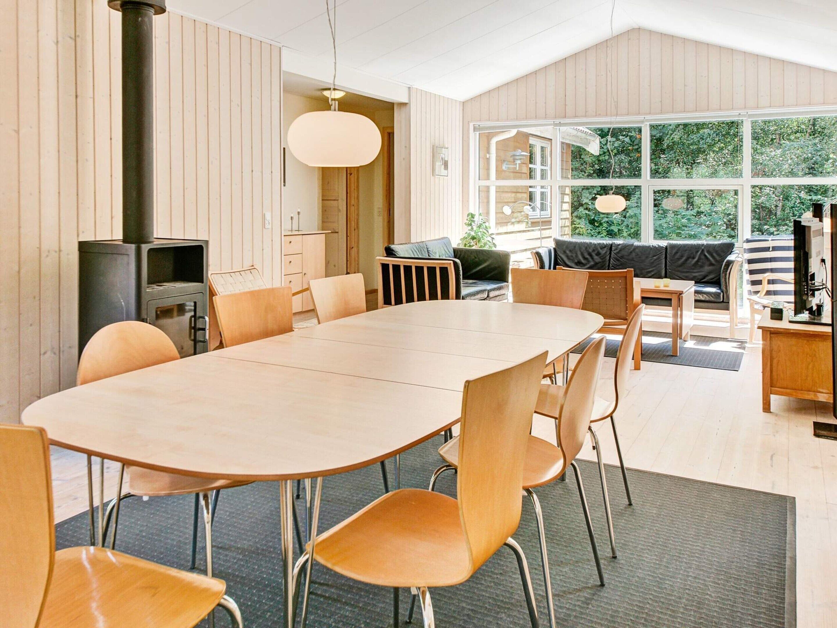 A modern interior with a large oval table, wooden chairs, a television, and a view of greenery through large windows.