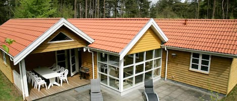 A two-story wooden house with a red-tiled roof, a patio with outdoor furniture, and a large window.
