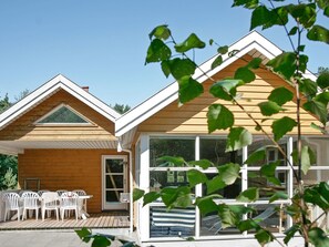 Ein Holzhaus mit überdachter Terrasse, weißen Stühlen und einem Fenster mit Blick auf einen Garten.