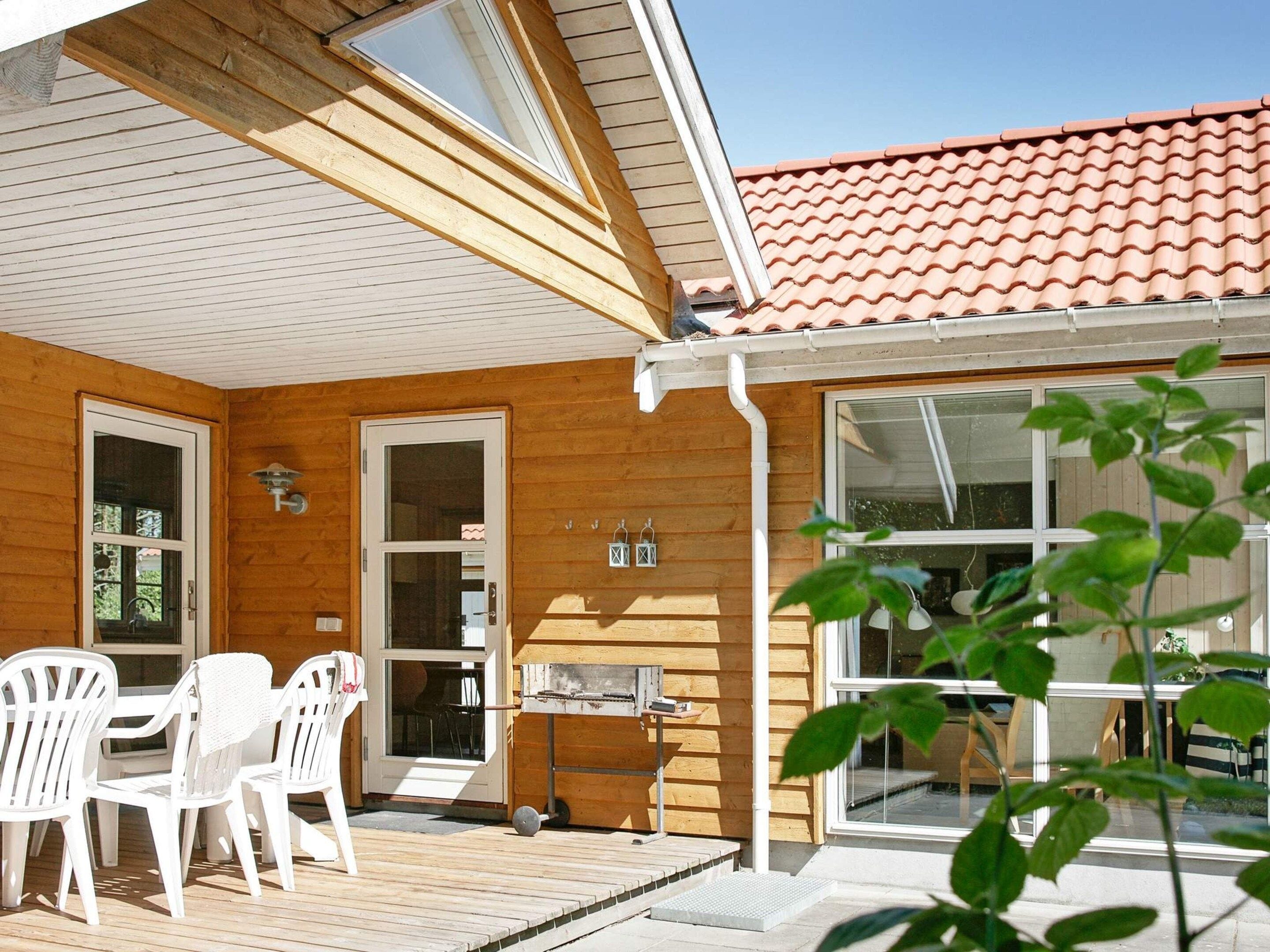 Une cabane en bois avec une terrasse, des chaises blanches, un barbecue et une fenêtre donnant sur la terrasse.