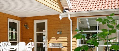 Une cabane en bois avec une terrasse, des chaises blanches, un barbecue et une fenêtre donnant sur la terrasse.