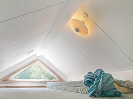 Una habitación con techo inclinado, tragaluces, cama con manta azul y lámpara fijada a la pared.
