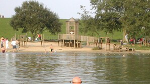 A lakeside park with a wooden play structure, picnic tables, and people enjoying the area.
