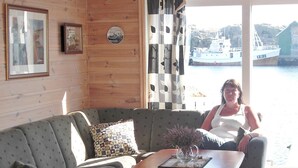 Een kamer met houten inrichting, een grijze bank, een gestreept kussen, een houten salontafel met een vaas, en uitzicht op een boot door een raam.