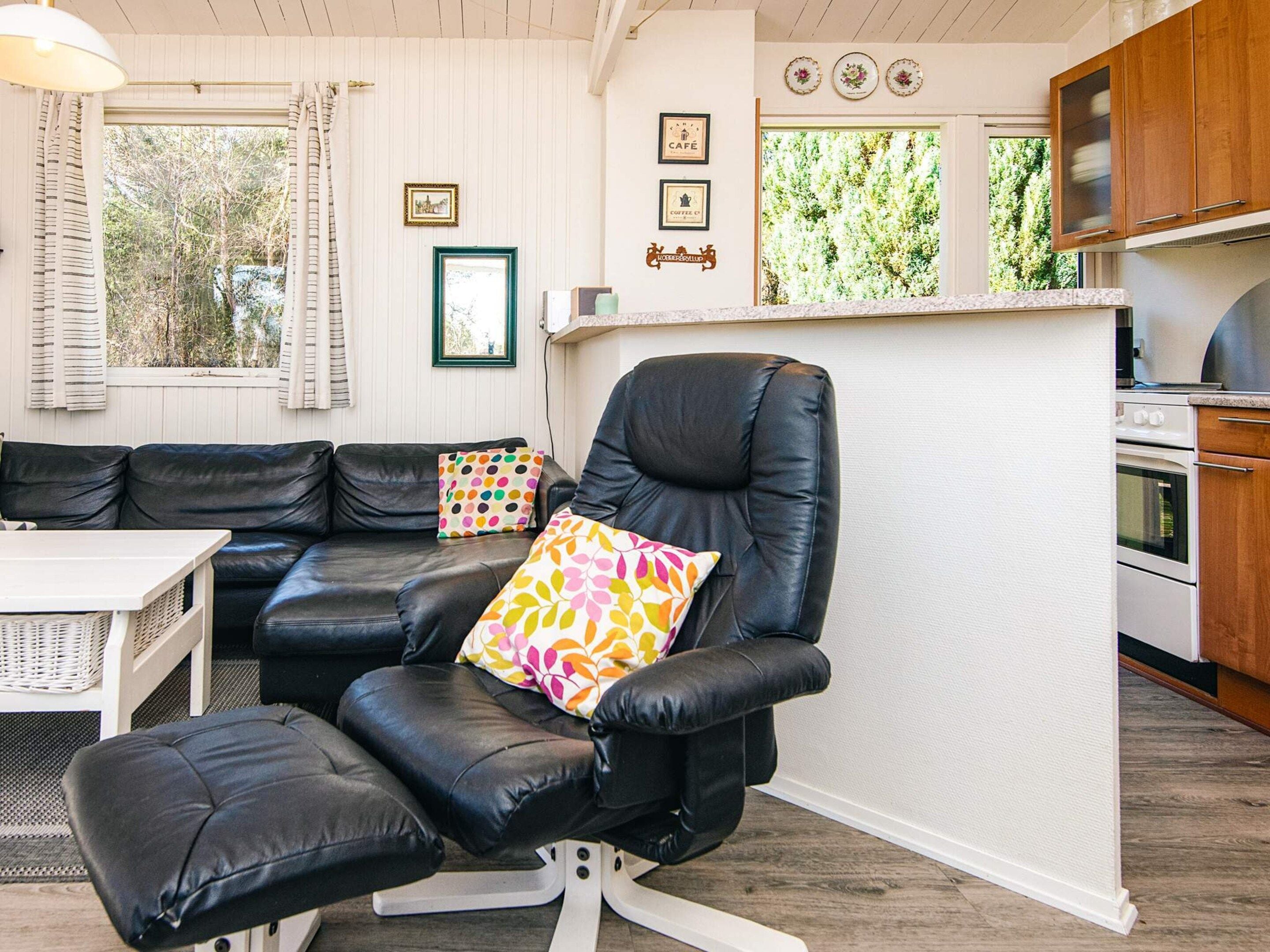 A cozy living room with a black leather sofa, a colorful pillow, and a white coffee table. There is a kitchen area with wooden cabinets and a white countertop.