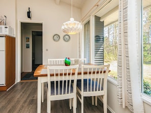 Un comedor con una mesa de madera y sillas blancas, un colgador de luces y vistas al exterior a través de una ventana con persianas.
