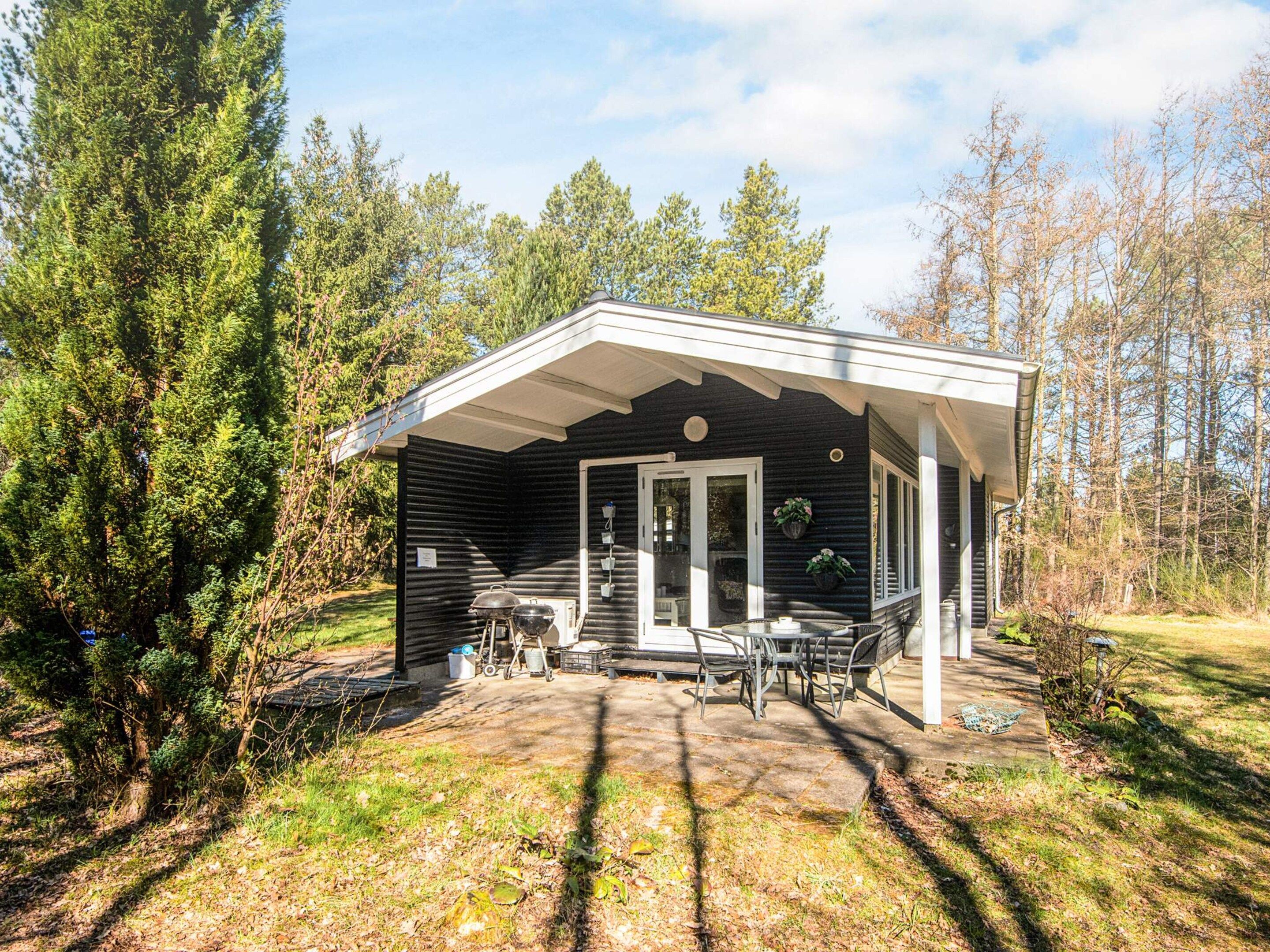 Ein kleines, eingeschossiges Blockhaus mit Veranda, Tisch und Stühlen, umgeben von Bäumen.