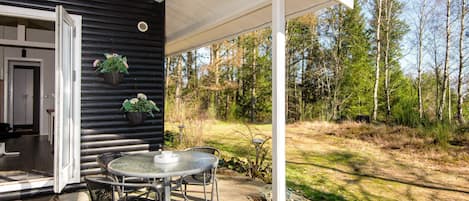 Een overdekt terras met een tafel en stoelen, een deur naar binnen en uitzicht op het omliggende bos.