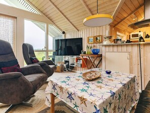 A cozy living room with a television, a dining table with a starfish tablecloth, and a large window overlooking a grassy area. - 5 Star Holiday Home in Hojslev (Hojslev)