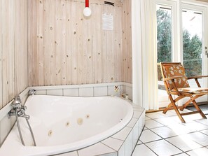 A bathroom with a large bathtub, wooden panel walls, and a chair near a sliding glass door. - 8 Person Holiday Home in Laeso (Læsø)