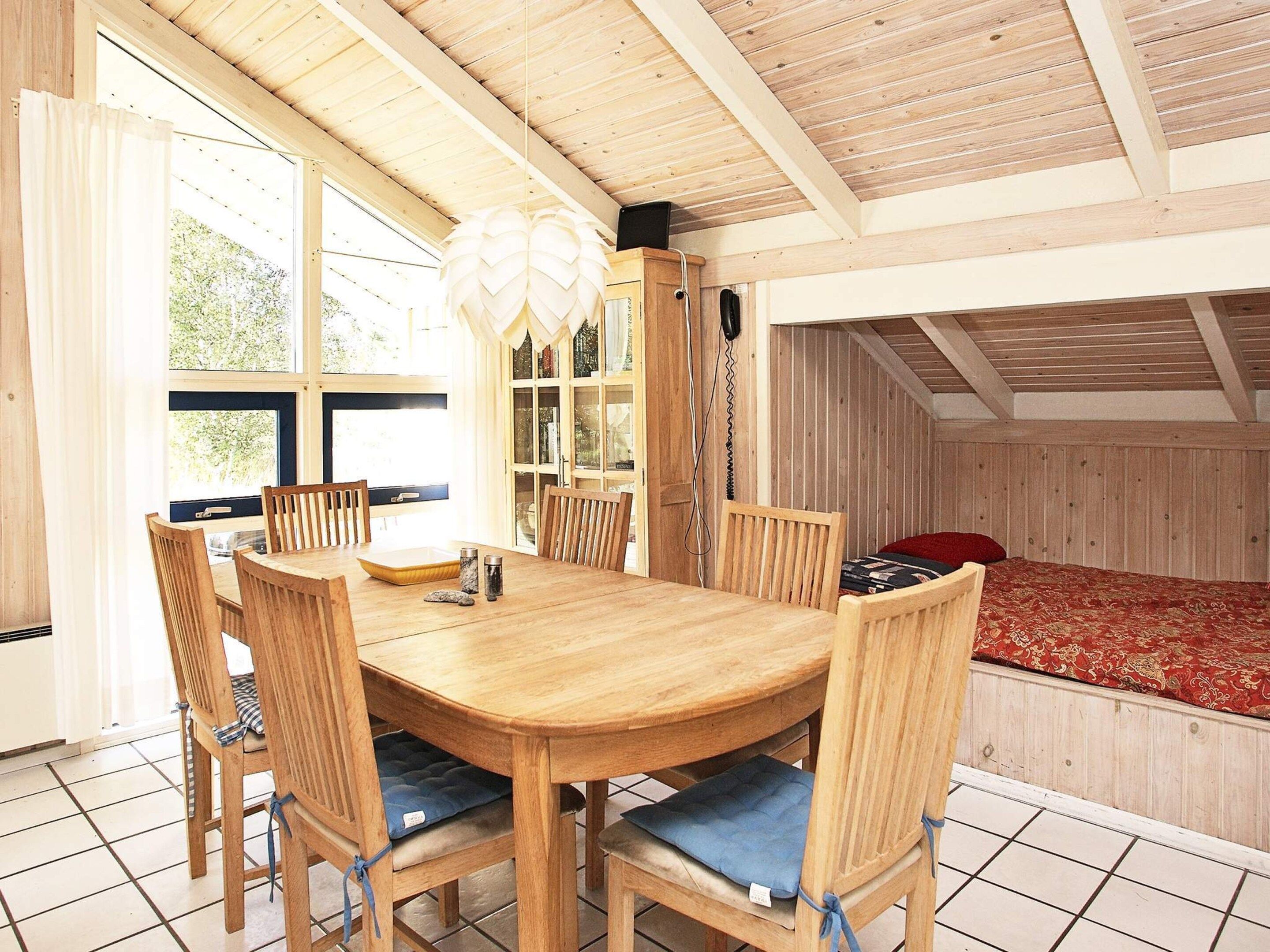 Une table à manger en bois, avec six chaises, un lustre et une vue sur l’extérieur grâce à de grandes fenêtres.