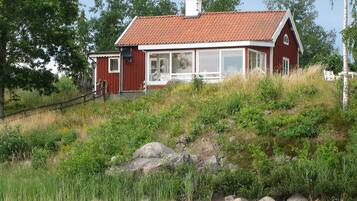 A red wooden house with a tiled roof, situated on a grassy hill by a body of water.