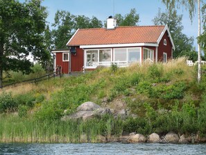 A red wooden house with a tiled roof, situated on a grassy hill by a body of water. - Holiday Home in Åtvidaberg (Atvidaberg)